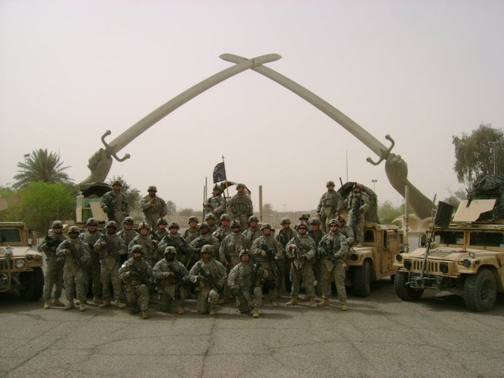 Military personnel gathered under a large decorative archway made of hooks, with military vehicles and desert landscape in background